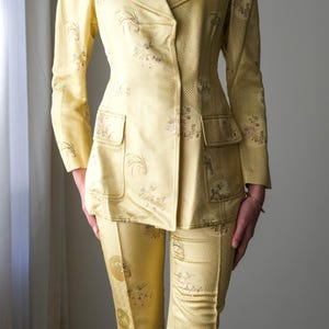 May include: A two-piece yellow suit with a floral pattern. The suit includes a blazer with a notched collar and two front pockets, and matching fitted pants. The fabric has a subtle geometric design.