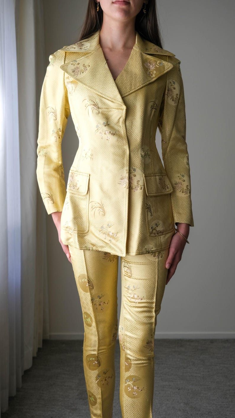 May include: A two-piece yellow suit with a floral pattern. The suit includes a blazer with a notched collar and two front pockets, and matching fitted pants. The fabric has a subtle geometric design.