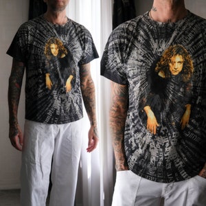 May include: Black graphic t-shirt featuring a portrait of a person with long brown hair and outstretched arms. The shirt has a radial design with text in a light gray color. The person is wearing white pants.