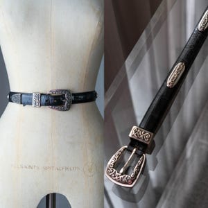 May include: Black leather belt with silver-tone metal buckle and accents. The belt has a textured pattern and decorative embellishments on the buckle and strap. The buckle features an ornate, vintage-inspired design.