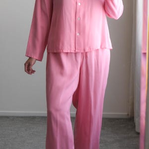 May include: A light pink pajama set. The set includes a long-sleeved button-down shirt and wide-leg pants. The shirt has a collar and the pants have a drawstring waist. The fabric appears to be a soft, lightweight material. The model is wearing light pink heeled sandals.