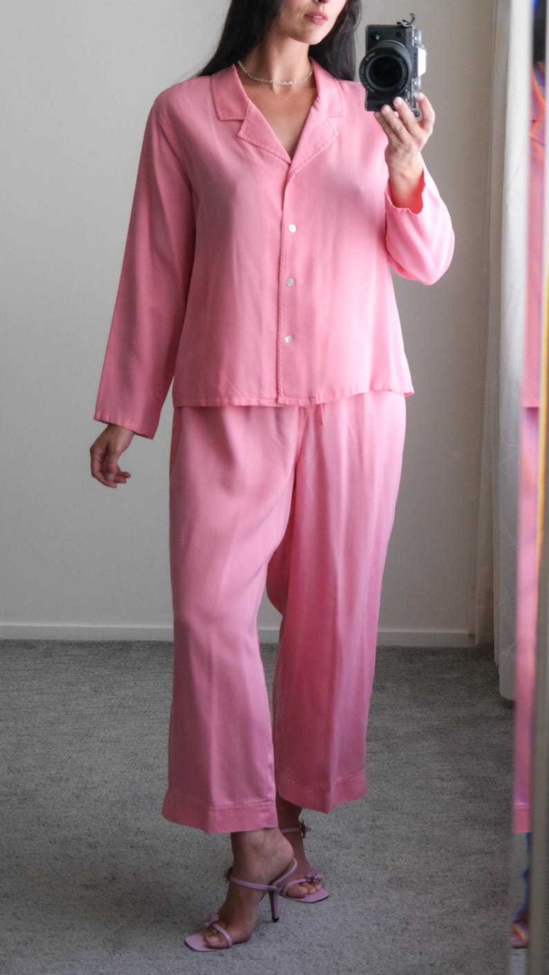 May include: A light pink pajama set. The set includes a long-sleeved button-down shirt and wide-leg pants. The shirt has a collar and the pants have a drawstring waist. The fabric appears to be a soft, lightweight material. The model is wearing light pink heeled sandals.