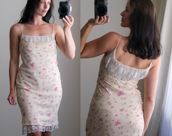 May include: A cream-colored slip dress with a pink floral pattern and delicate spaghetti straps. The dress features a lace trim at the neckline and hem. The dress is approximately knee-length.