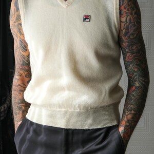 Peut inclure: Gilet en tricot cr&egrave;me &agrave; col en V avec un petit &eacute;cusson logo. Le gilet est associ&eacute; &agrave; un pantalon en satin noir. L'ensemble est de style classique, adapt&eacute; au superposition. Le gilet a un ourlet et un col c&ocirc;tel&eacute;s.