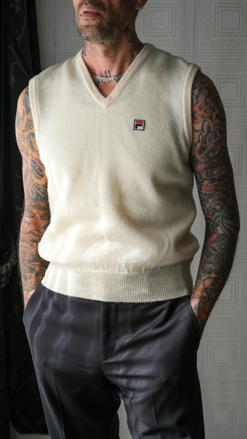 Peut inclure: Gilet en tricot cr&egrave;me &agrave; col en V avec un petit &eacute;cusson logo. Le gilet est associ&eacute; &agrave; un pantalon en satin noir. L'ensemble est de style classique, adapt&eacute; au superposition. Le gilet a un ourlet et un col c&ocirc;tel&eacute;s.