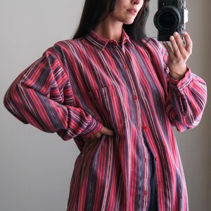 May include: A button-down shirt with vertical stripes in shades of red, purple, and white. The shirt has a single chest pocket and long sleeves. The model is wearing the shirt with dark blue pants.