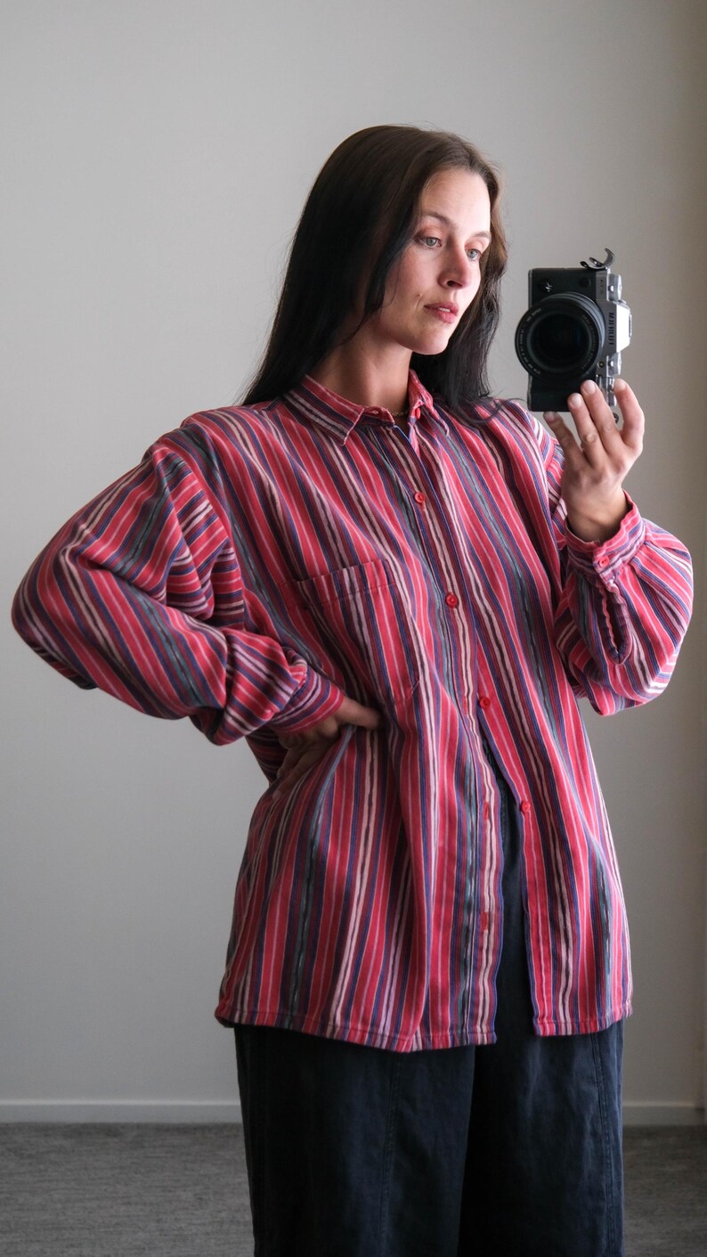 May include: A button-down shirt with vertical stripes in shades of red, purple, and white. The shirt has a single chest pocket and long sleeves. The model is wearing the shirt with dark blue pants.