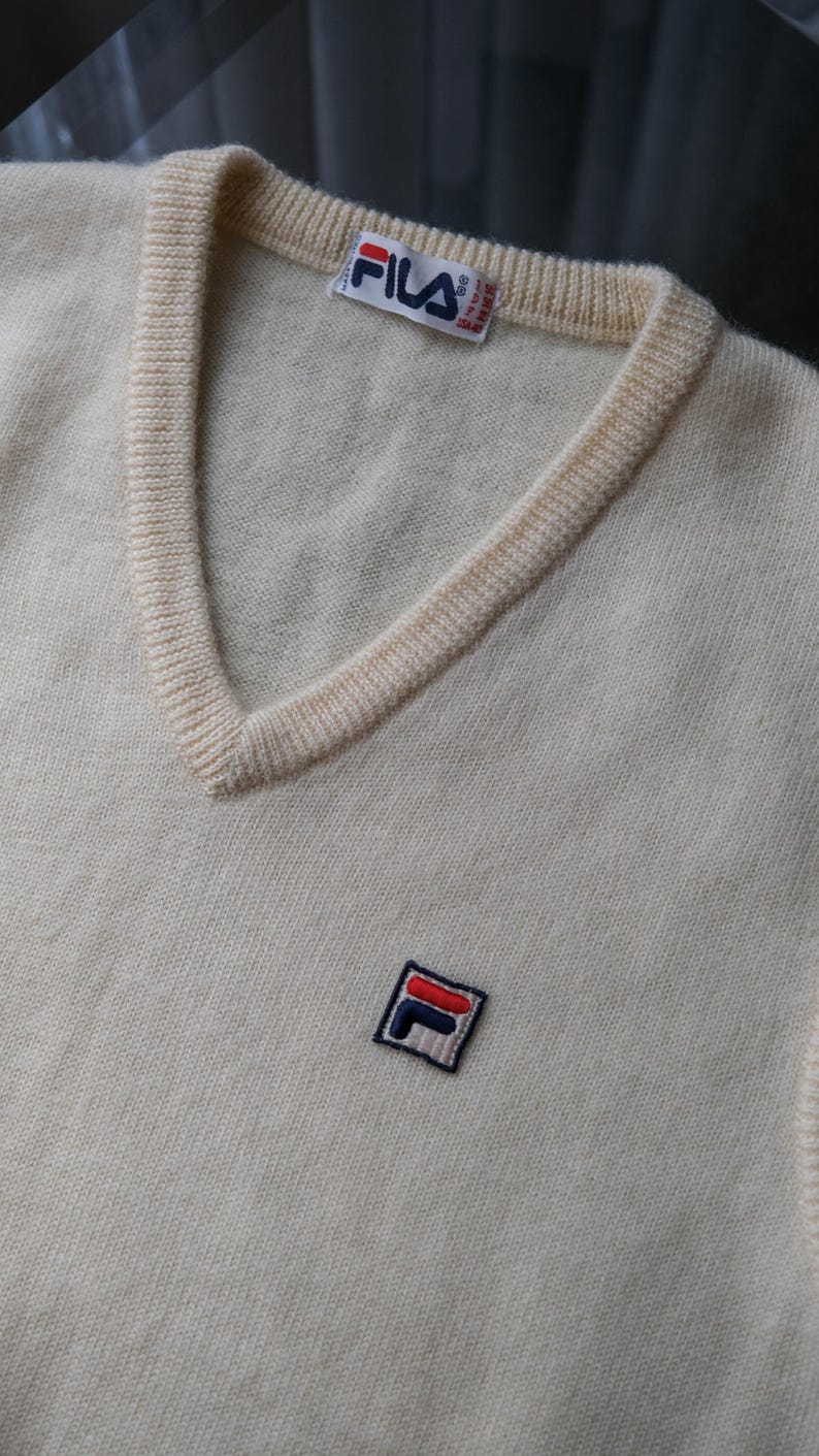 Peut inclure: Pull-over cr&egrave;me &agrave; col en V avec un petit &eacute;cusson FILA sur la poitrine. Le pull a une encolure c&ocirc;tel&eacute;e et une petite &eacute;tiquette FILA. Le pull est fait d'une mati&egrave;re tricot&eacute;e douce.