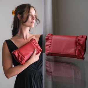 May include: A rectangular red clutch bag featuring a smooth central panel and gathered, ruffled fabric detailing along both side edges. The accessory has a structured flap closure and a consistent deep red colour throughout its exterior surface.