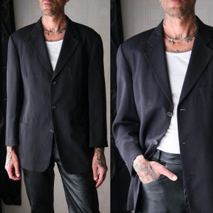 Op de afbeelding: Een zwarte blazer met een sluiting met drie knopen. De blazer wordt gecombineerd met een wit ondershirt en een zwarte broek. De jas heeft een klassieke revers en een getailleerde pasvorm, geschikt voor formele of semi-formele gelegenheden.