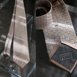May include: A light brown silk tie with a repeating pattern of small, light blue and white floral designs. The tie is displayed on a clear stand, with a close-up showing the label "GIANFRANCO FERRE".