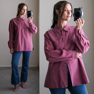 May include: A dusty rose button-down shirt with long sleeves and a chest pocket. The shirt is paired with blue denim jeans. The model is holding a camera.