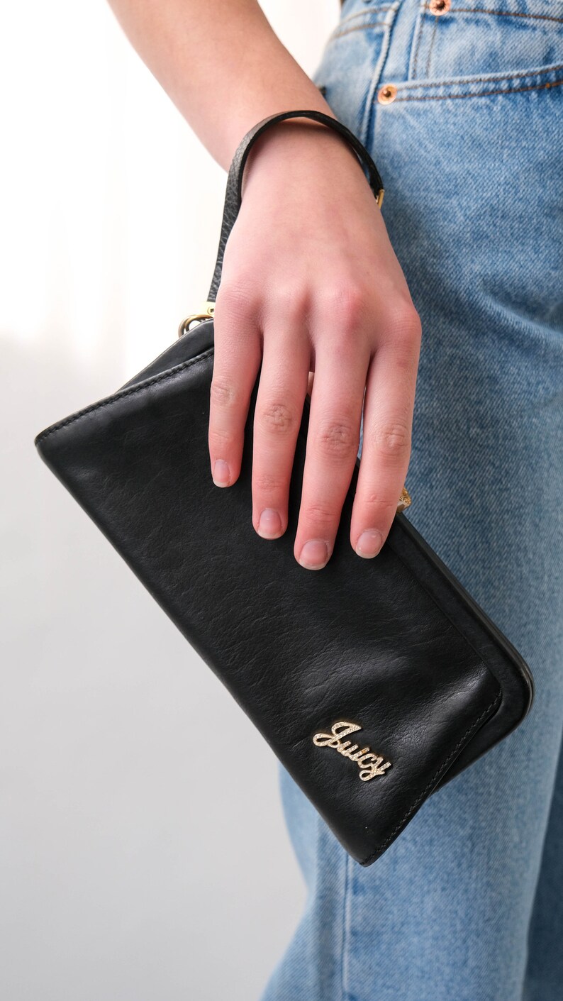 May include: A black leather wallet with a gold Juicy Couture logo. The wallet is being held by a hand.