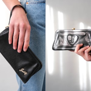 Może przedstawiać: Czarna skórzana kopertówka z złotym logo Juicy Couture i złotym sercem z literą J w środku. Kopertówka ma pasek na nadgarstek.