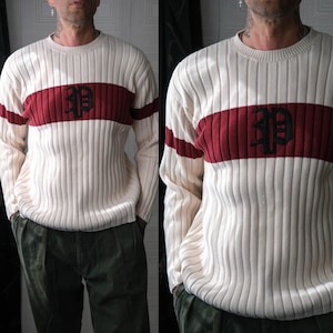 May include: Cream-colored ribbed sweater with a burgundy stripe across the chest and sleeves. The sweater features a black embroidered design on the burgundy stripe. The long-sleeved sweater is a classic style.