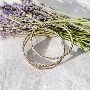 May include: Three silver bangle bracelets with a twisted design, arranged on a white fabric background with a bouquet of lavender flowers.