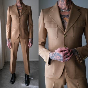 Könnte beinhalten: Ein beigefarbener Anzug mit einer Jacke mit vier Taschen und einer passenden Hose. Die Jacke hat ein fallendes Revers und goldfarbene Knöpfe. Der Anzug ist aus einem glatten Stoff gefertigt und hat eine klassische, strukturierte Passform.