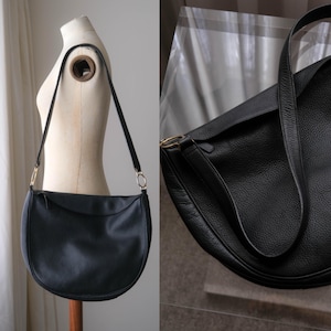 May include: A black crescent-shaped shoulder bag with a pebbled texture, featuring a single long strap attached with gold-toned metal rings. The bag has a curved bottom edge and a top zipper closure, displayed on a mannequin form for shape.