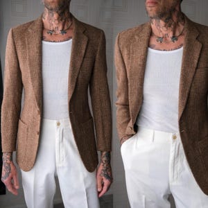 Könnte beinhalten: Ein brauner Blazer mit dezentem Karomuster, kombiniert mit einem weißen Unterhemd und einer weißen Hose. Der Blazer hat ein klassisches Zwei-Knopf-Design und zwei Vordertaschen. Die Hose hat einen Knopfverschluss.