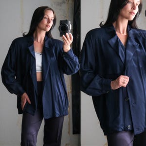 May include: A navy blue blazer with a classic collar and button closure. The jacket has long sleeves and a slightly oversized fit. The fabric has a subtle sheen. The blazer is paired with dark trousers.