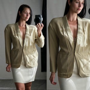 May include: A light beige blazer with a classic lapel and two front pockets. The blazer is paired with a white skirt. The outfit is a neutral color, suitable for a variety of occasions. The blazer has a single button closure.