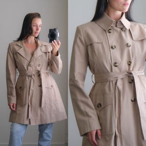 May include: A beige double-breasted trench coat featuring a collared neckline, long sleeves, and a matching fabric tie belt at the waist. The coat includes two front flap pockets and dark brown buttons arranged in two vertical rows down the front.