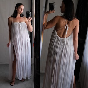 Op de afbeelding: Een lange, witte jurk met dunne bandjes en een hoge split. De jurk heeft een los, vloeiend ontwerp met een getextureerde uitstraling. Kwastjes sieren de bandjes en de achterkant van de jurk. De jurk is gemaakt van een lichte stof.