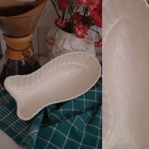 Puede incluir: Una fuente de cerámica color crema con forma de pez, que presenta un patrón de escamas texturizado en la superficie interior y un borde festoneado. El plato ovalado alargado tiene una cola y una cabeza definidas, con un acabado liso y brillante.