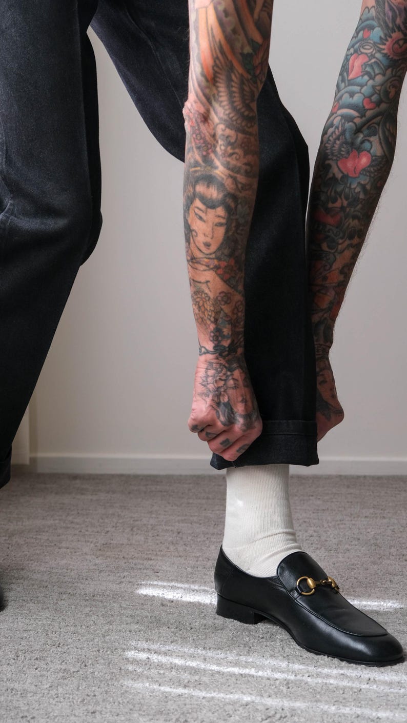 Peut inclure: Mocassins en cuir noir avec boucle dor&eacute;e. Les chaussures sont port&eacute;es avec des chaussettes blanches et un jean denim fonc&eacute;. L'image met l'accent sur les chaussures et le bas des jambes.