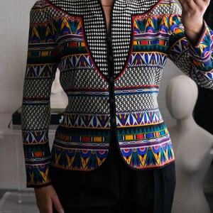 May include: A black and white patterned blazer with colorful geometric designs. The blazer has a mandarin collar and a button closure. The model is wearing black pants.