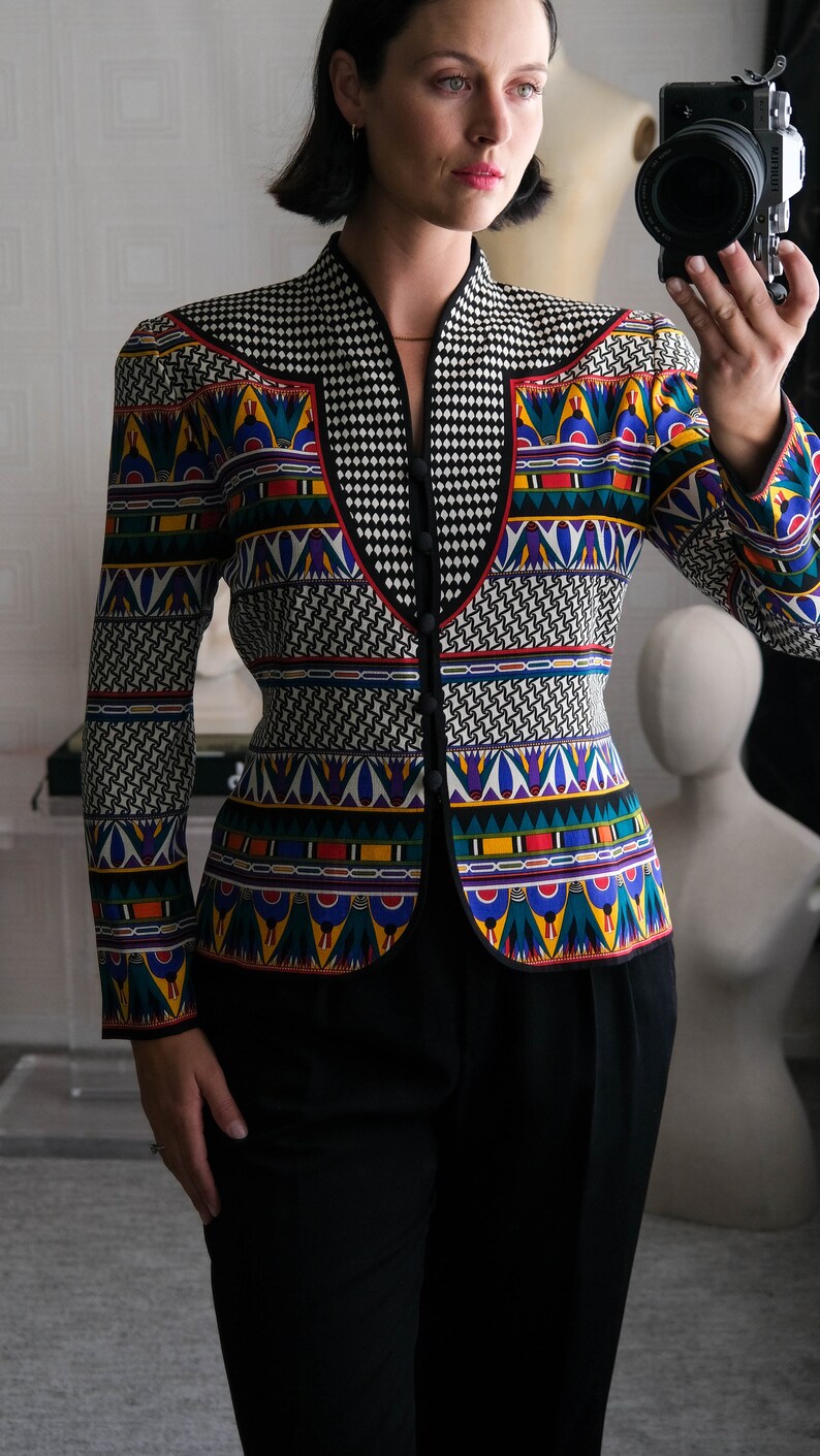May include: A black and white patterned blazer with colorful geometric designs. The blazer has a mandarin collar and a button closure. The model is wearing black pants.