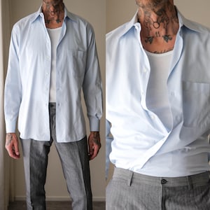 May include: A light blue button-down shirt with a chest pocket. The shirt is paired with gray trousers. The shirt is unbuttoned, revealing a white undershirt. The sleeves are buttoned at the cuffs.