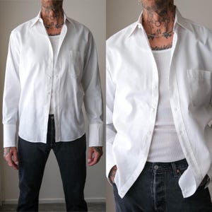 May include: A crisp white long-sleeve button-down shirt featuring a classic collar, a single chest pocket, and wide cuffs. The shirt is worn unbuttoned over a white ribbed tank top, paired with dark charcoal denim jeans, creating a layered look.
