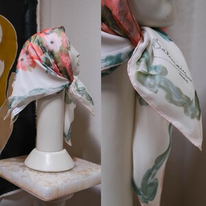 May include: A white silk scarf with a floral pattern in shades of pink, green, and white. The scarf is draped over a mannequin head. The scarf has a pink border and the words "Balmain Paris" are printed on the scarf.