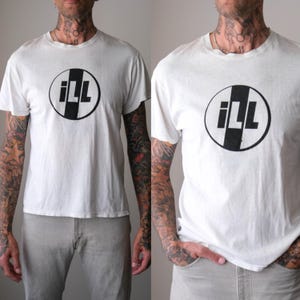 Puede incluir: Una camiseta blanca de manga corta con un gráfico circular negro en el pecho. El gráfico contiene una gruesa franja vertical negra que divide el círculo, con las letras iLL impresas en una fuente negrita sin remates en el centro.