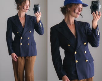 Puede incluir: Un blazer azul marino de doble botonadura con botones dorados en la parte delantera y en los puños, combinado con pantalones marrones. En una vista, se lleva una gorra de béisbol azul con texto blanco bordado. El blazer tiene una solapa clásica.