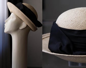 May include: A beige straw hat with a black ribbon wrapped around the crown. The hat is displayed on a mannequin head. The brim is slightly curved, and the ribbon adds a touch of elegance. The hat is a classic accessory.