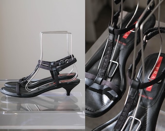 Puede incluir: Sandalias negras Prada con diseño de tira en T, con tiras de tela finas, un tacón pequeño y una etiqueta con el logo rojo en la plantilla. Las sandalias se muestran sobre soportes metálicos, exhibiendo la marca Prada y el texto Made in Italy.