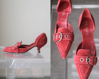 Puede incluir: Un par de zapatos de tacón bajo en ante rojo con punta afilada, aberturas laterales, una tira decorativa sobre el empeine con una hebilla de metal plateado y costuras blancas en contraste. El logotipo de Prada aparece en la plantilla interior.