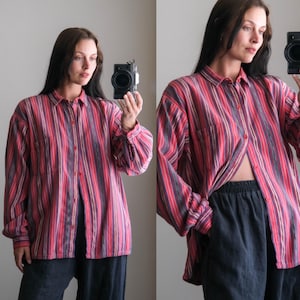 May include: A button-down shirt with vertical stripes in shades of red, pink, purple, and gray. The long-sleeved shirt has a chest pocket and is worn open. The shirt is paired with dark blue pants.
