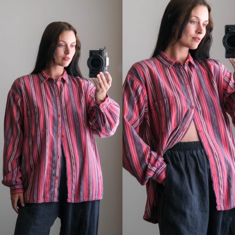 May include: A button-down shirt with vertical stripes in shades of red, pink, purple, and gray. The long-sleeved shirt has a chest pocket and is worn open. The shirt is paired with dark blue pants.