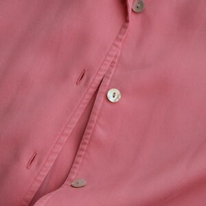 May include: Close-up of a pink button-down shirt. The shirt features a classic collar and several pearlescent buttons. The fabric has a subtle texture, and the overall aesthetic is clean and minimalist.