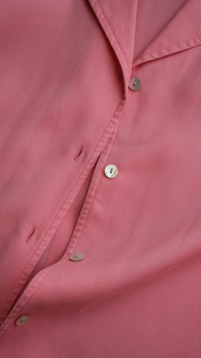 May include: Close-up of a pink button-down shirt. The shirt features a classic collar and several pearlescent buttons. The fabric has a subtle texture, and the overall aesthetic is clean and minimalist.