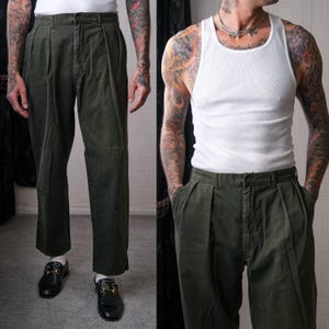 Puede incluir: Pantalones plisados ​​verde oliva con un ajuste relajado. Los pantalones tienen cintura alta y pernera recta. Los pantalones se combinan con mocasines de cuero negro con detalles dorados. El modelo lleva una camiseta sin mangas blanca.