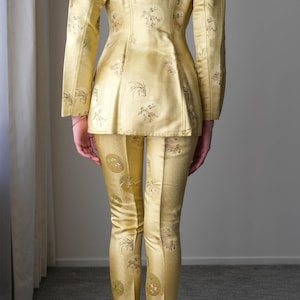 May include: A golden yellow suit with a floral pattern. The suit includes a fitted jacket and matching pants. The person is wearing pink heels. The fabric has a subtle sheen, suggesting a formal or vintage style.