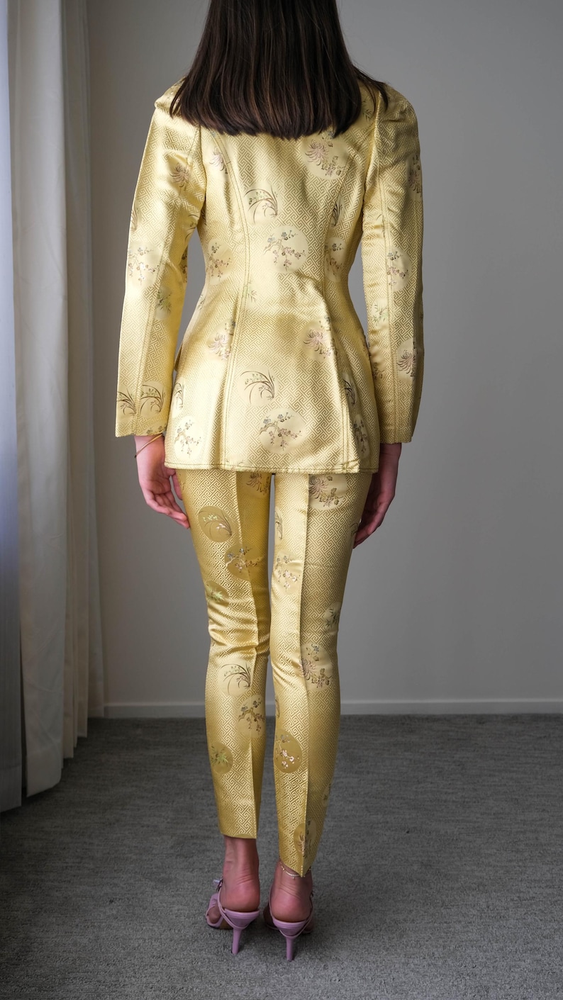 May include: A golden yellow suit with a floral pattern. The suit includes a fitted jacket and matching pants. The person is wearing pink heels. The fabric has a subtle sheen, suggesting a formal or vintage style.