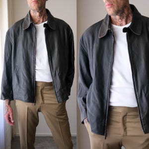 Puede incluir: Una chaqueta con cremallera en color gris carbón oscuro con cuello clásico, usada sobre una camiseta blanca lisa y combinada con pantalones color canela. La chaqueta presenta una textura lisa en los paneles frontales y un patrón sutilmente acanalado en las mangas.