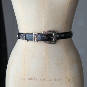 May include: A black belt with a textured surface and silver-coloured metal buckle and accents. The buckle has an ornate design. The belt is displayed on a mannequin.