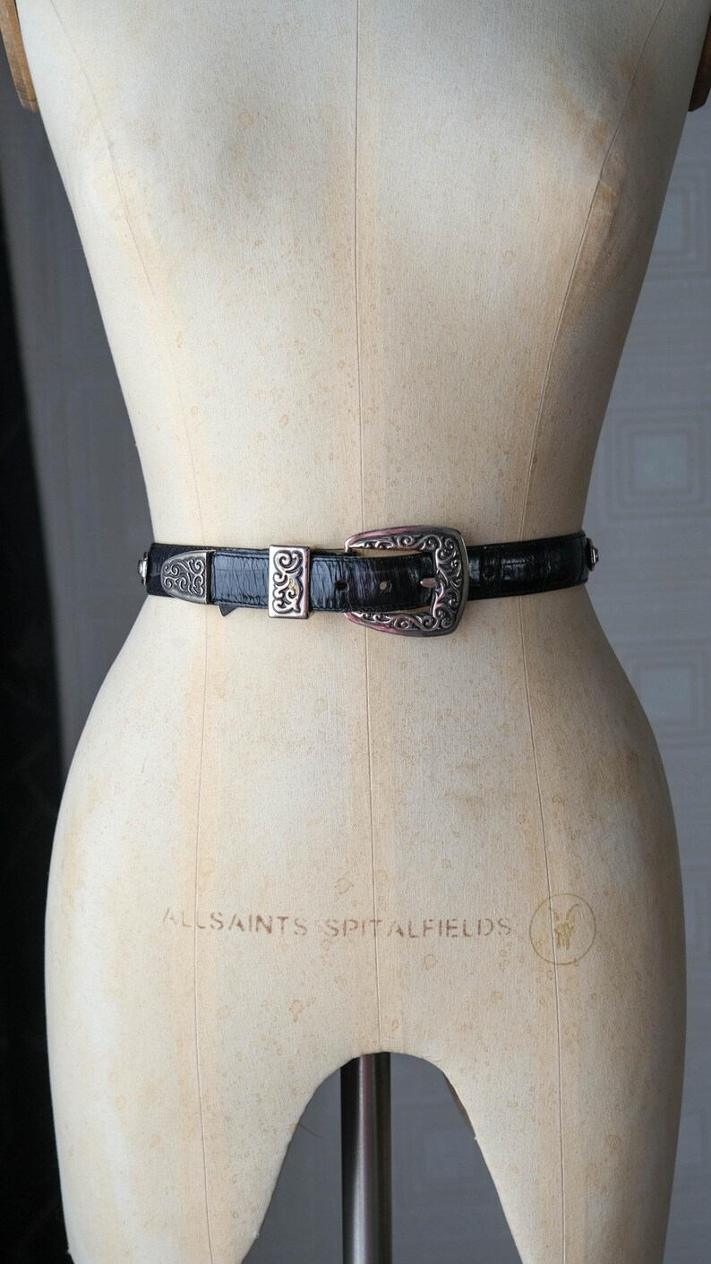 May include: A black belt with a textured surface and silver-coloured metal buckle and accents. The buckle has an ornate design. The belt is displayed on a mannequin.