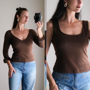 May include: A brown long-sleeve top with a deep V-neck and fur cuffs, paired with light blue jeans. Next to it, a brown tank top with a scoop neck. The tops are made of a soft material. The image is a mirror selfie.
